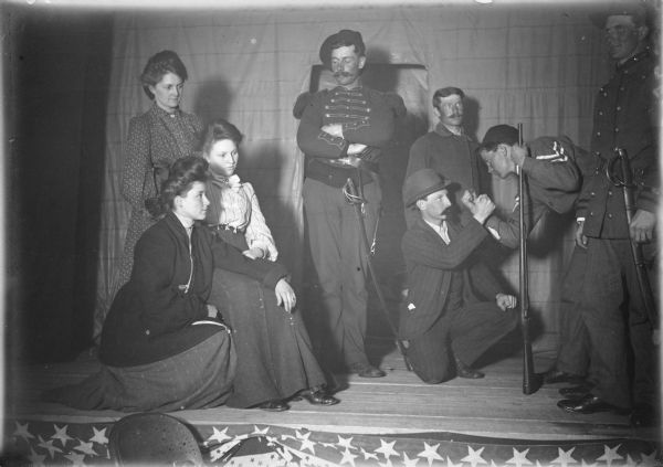 Three young women and five young men, three of whom are in military uniform, are posing in a scene from a play. There is patriotic bunting hanging from the front of the small stage.