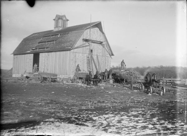 Will von der Sump, far right, is standing near the belt leading from a portable steam engine to a shredder and elevator next to a barn with a cupola ventilator. Another man is standing atop a wagonload of corn stalks as two men are feeding them into the shredder. Two horses are hitched to a wagon nearly full of ears of corn; a man is sitting at the rear of the wagon. Another man is standing in the door of the barn at left.