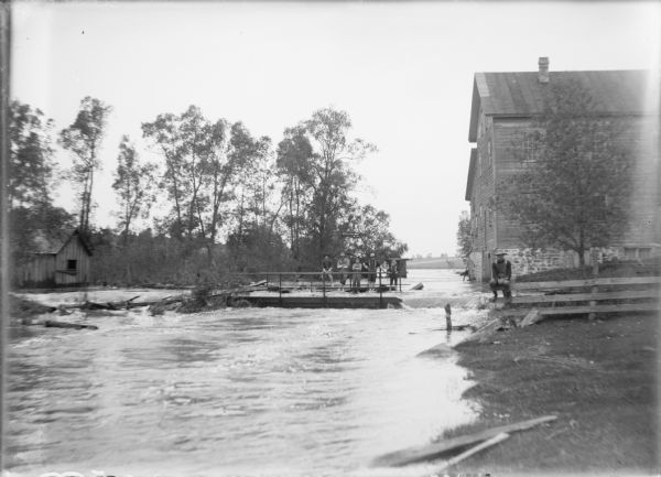 View upstream towards five young men and boys posing on the flooded bridge at Dates Mill as water rushes from the swollen mill pond into French Creek. Another young man is sitting on a fence at right. Behind him is the mill, and there is a small building at far left.