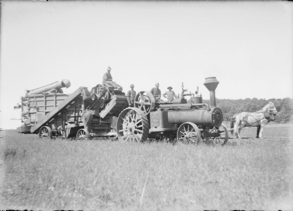 Three men are posing on a large steam tractor, and a fourth man is sitting on a threshing machine behind the tractor. The large drive belt is coiled and is hanging near the front of the thresher. Two horses in harness are standing at far right. Written on the negative envelope: "Hamilton threshing outfit."