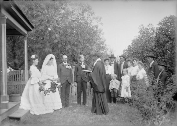 A group of young adults and children are posing outdoors near the open porch of a house in a mock wedding ceremony. The groom, third from left, is portrayed by August Bernhagen. The bride and her attendant are at left near the porch; the best man is standing to the right of the groom. The officiant is holding a book and is wearing a cassock. Members of the congregation at right include Mae von der Sump, fifth from right. A note on the negative envelope states: "Mock Marriage at Uncle's."
