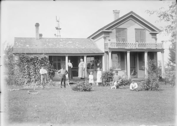 A group is posing in front of a two-story wood frame house with a lower wing at left. Mae von der Sump is standing near a column at the left of steps leading onto a porch. An unidentified older woman is leaning against the column at right. Two girls in light-colored dresses are standing on the lawn. There are two men standing at left near a push lawn mower. A man at right is posing with a dog, and a young man is sitting on the lawn nearby. A windmill is behind the house and there is a dinner bell mounted on the windmill's tower. Vines shade the left end of the porch.