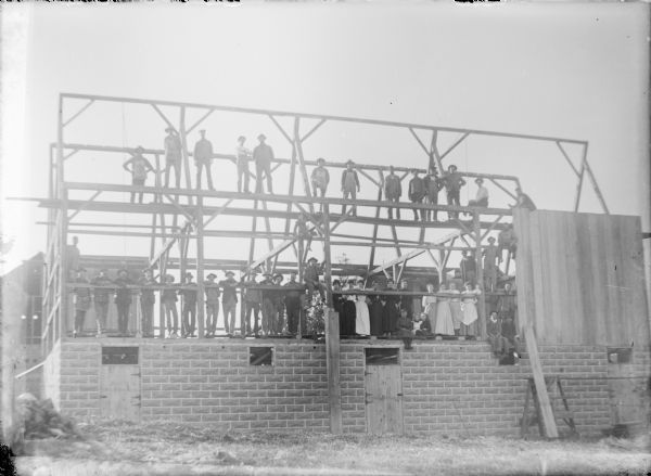 A crowd of men, women and children are posing on the beams and floor of a new barn on the Frank von der Sump farm. The barn has a concrete block foundation. A portion of the barn siding has been applied at right.