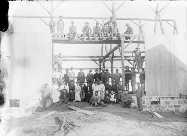 A crowd of men, women and children are posing on the beams and floor of a new barn on the Frank von der Sump farm. The barn has a stone foundation. A portion of the barn siding has been applied. Frank von der Sump is sitting on the ground in the center of the doorway, and is holding a small dog beside him. Sitting directly behind him is his daughter Leta. Several of the men are holding hammers.