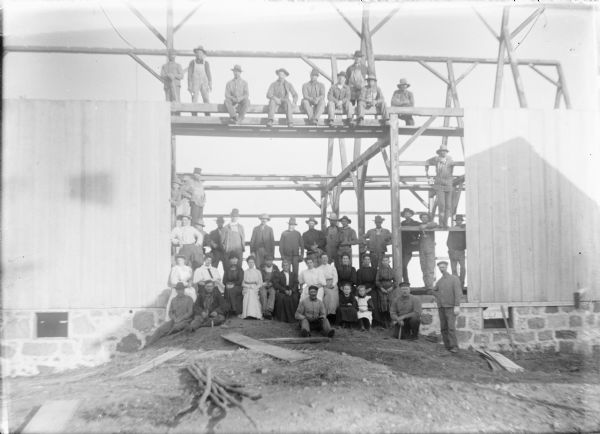 A crowd of men, women and children are posing on the beams and floor of a new barn on the Frank von der Sump farm. The barn has a stone foundation. A portion of the barn siding has been applied. Frank von der Sump is sitting on the ground in the center of the doorway. Sitting directly behind him is his daughter Leta. Several of the men are holding hammers.