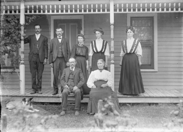 An older man and woman are sitting on chairs in front of a low porch. Behind them, on the porch, two men and three women are standing. The two women at right are dressed alike, with similar skirts and matching light-colored blouses with dark ribbon trim. There is a dog lying on the grass at left.