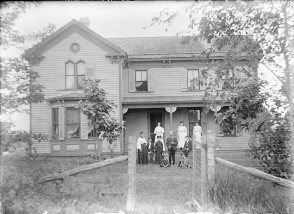 Members of the James Kearns family are photographed through the open gate of a woven wire fence. They are posing on and in front of the porch of a large two-story frame house. The house has painted trim, shutters and decorative eave brackets. An unidentified elderly woman is standing in the center of the group; James is to the right and his wife Clara is at far left. Their sons James, left, and Edward are sitting on the porch floor. John (partially obscured by a dog) and Thomas are sitting on the lawn. Daughters Mary, Winnie and Annie (not necessarily in order) are standing on the porch.