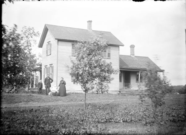 View across lawn towards a well-kept, two-story wood frame house with two visible one-story extensions, both with a porch. On the left a man and woman are posing with an infant in a baby carriage, and an older boy is standing to the left of the man. There is a dog in front of the carriage.