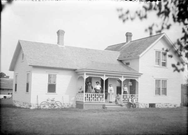 Valentine Kusick is sitting in the grass in a chair in front of the porch railing of his well-kept house. His wife Elizabeth is sitting at the top of the porch steps. On the porch are their children, (not necessarily in order) Eliza, Agnes, John and Joseph. The one and one-half story house has a single story wing on the left. There is a bicycle leaning against the side of the house between two rifles. In the background on the left is a barn.