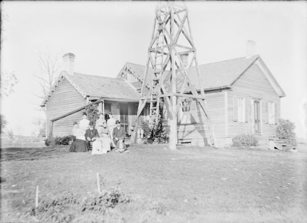 A wooden windmill tower dominates the front yard of a small one-story wood frame house. A group of people are posing on the left, including two couples sitting on chairs. The woman sitting at far left is holding an infant in her arms. Three women are standing behind them, and a girl and boy are sitting on the ground in front. There are buckets on the porch.