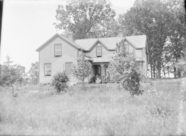 View over tall grass towards Ed Kearns, standing at far right, posing with his family near the porch of the house. A little girl is standing next to him, holding his pant leg. His wife is sitting on the porch floor holding an infant; two boys wearing suits are standing beside her. The house is a one and one-half story wood frame structure. There are young trees in the front yard.