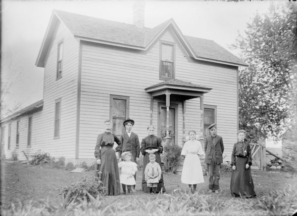 A group of people identified only as "Mrs. Ed Wallace Family" are posing on the lawn of a one and one-half story wood frame house. A little boy in a sailor outfit and girl in a short light-colored dress are standing in front at left. Behind them are two women in dark dresses standing on either side of a young man wearing a suit and tie. To the right are a young woman in a light-colored dress with a large bow in her hair standing next to a young man in work clothes. At far right is a woman in a dark dress.