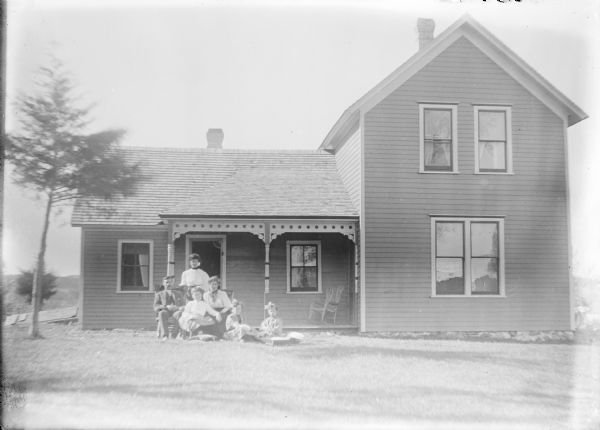 View across lawn towards the John McLane family posing in front of the porch of their well-kept one and one-half story wood frame house. Family members include John and his wife Marietta sitting with their daughter Margarette between them. Daughter Ellen is standing behind them, and twins Ina and Ada are sitting on the lawn at right. There is a wicker rocking chair on the porch.