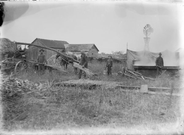 A portable horse-driven sorghum mill, owned by the Cuff family, has been set up at the August Hernkind farm. Two horses are hitched to the tree trunk that turns the mill. Two men at left work to feed cane into the rollers, and a boy, center, is holding a bucket. A man at right is standing at the steaming evaporator pan. There are farm buildings and a windmill in the background.