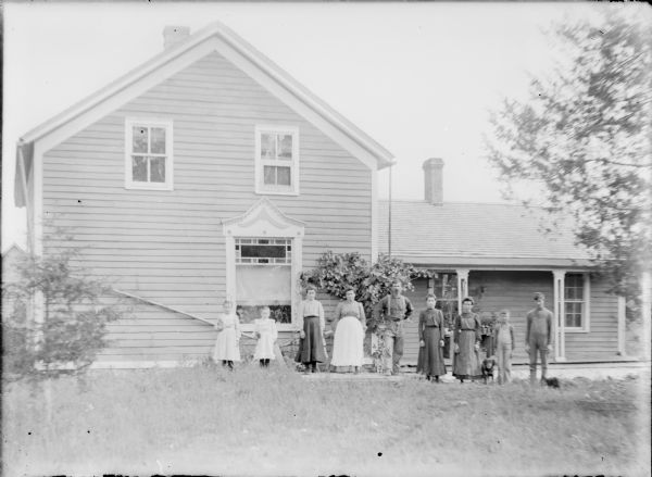 August Hernkind, center, and his wife Kate are posing with their seven children in front of their one and one-half story wood frame farm house. Their daughter Mary is standing at far left next to Nellie, the youngest. At far right is Henry, with George next to him. The older three girls, not necessarily in order, are Louise, Anna, and Katie. There is a dog next to George, and a cat appears to have been approaching the group from far right during the exposure.