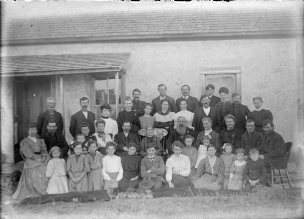 Jim Cheney, with a full beard, is sitting next to his wife Mary, left, surrounded by men, women and children, some sitting on rugs spread on the lawn in front of the Cheney's farm house. In the front row at left are twins Ina and Ada McLane. Their sister Margarette is to the right. Their mother, Marietta (Mrs. John) McLane, is sitting behind Margarette. Ellen McLane, wearing a light-colored blouse and dark collar is in the back row next to her father John.