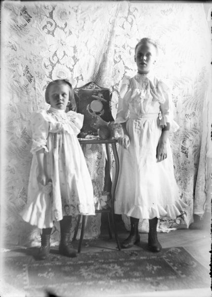 An available light, interior view of two girls, identified on the negative envelope as Ella and Thresa [sic] Alson, flanking a small table. There is a live squirrel on the table, eating a nut in front of a velvet covered photo album on a stand. The younger girl at left is holding a doll. The girls are wearing light-colored dresses and are standing in front of a lace drapery.
