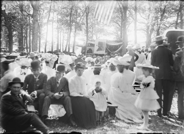 A crowd of men, women and children have gathered in a clearing in a wooded area for a July 4 celebration. Sunlight passing through an American flag highlights printing on the flag which reads: "Wm. Atkinson. Post N. 267. Below the flag there is patriotic bunting, and a caricature of Abraham Lincoln on an easel. The men are dressed in suits and the women are wearing large hats and light-colored blouses and dresses.
