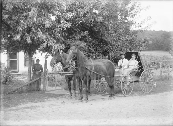 A young man, identified on the negative envelope as Arther Samfle [sic], is sitting with a young woman in a buggy hitched to a matched team. The horses are fitted with fly nets. A woman is standing behind a woven wire fence and gate near the horses. There is a house behind her.