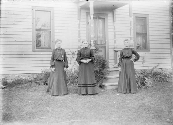 Three unidentified women wearing dark-colored dresses are posing standing in front of a wood frame house. Steps behind them lead to a small covered porch.