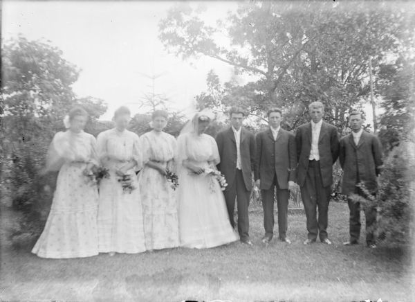Outdoor group portrait of a bride and groom, center, flanked by their six attendants, taken on the lawn of the Von der Sump farmhouse. Although not identified by the photographer, the bride appears to be Hattie Johanna von der Sump, who married Ellis Owens June 27, 1906. Leta von der Sump is standing next to the bride, her sister. Their brother Will von der Sump is second from right. The women are wearing long light-colored dresses and are holding small bouquets.