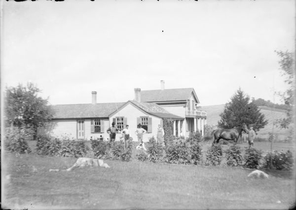 Three men and a woman are posing with a dog at the side of a rambling wood frame farm house. The house has a two-story section with a porch and two single-story extensions, one to the side with another to the rear. There is a fourth man with a horse on the front lawn.