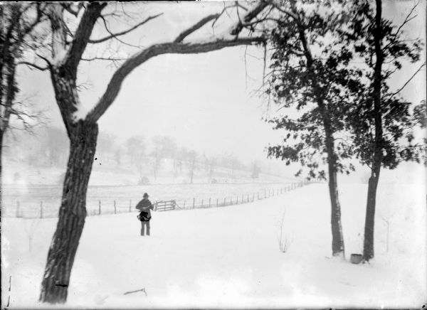 An unidentified man is posing with a long gun in a snowy field. There is a tree in the left foreground, and a wooded hillside in the background.