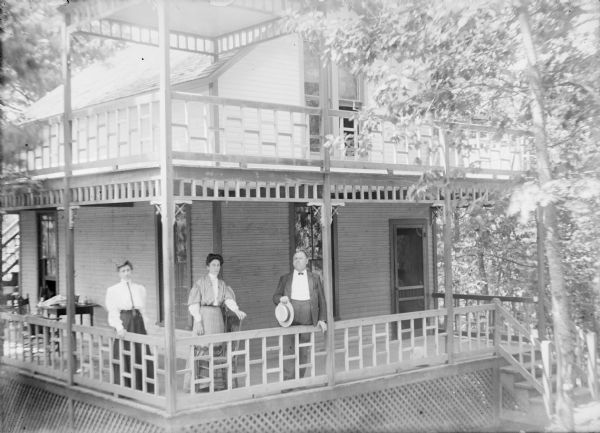 Slightly elevated view of an unidentified man and two women posing standing on the open porch of a two-story wood frame cottage at Wisconsin Dells. The porch also has an upper level, with a roof over the corner portion. There is decorative fretwork on the porch and lattice skirting underneath. The wood siding on the lower level of the cottage is unpainted.