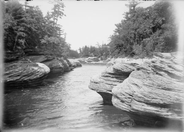 A summer view of rock formations known as the Navy Yard lining the Wisconsin River at the Dells. Further down the river is the Larks Hotel, with the hotel's name on it on the roof. Boats are along the shoreline near a long, wooden stairway that leads up the rocks from the river to the hotel.