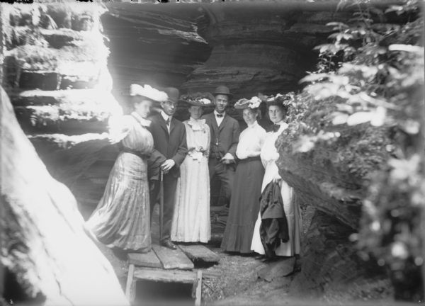 A group of two men and four women, including Hattie, far left, and Leta von der Sump, far right. The well-dressed group is posing in a narrow passage created by rock formations. The three people at left are standing on a plank bridge over a small stream.