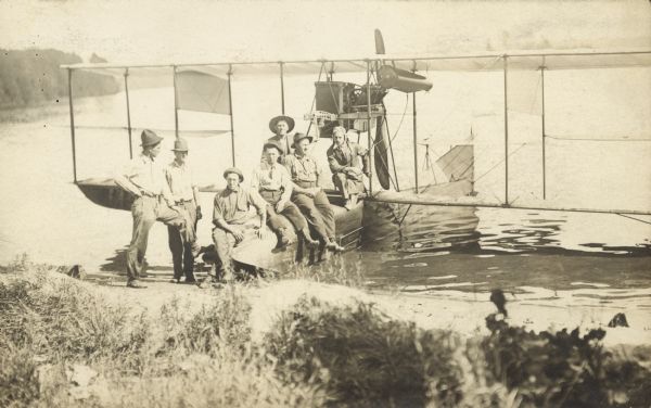 View from shoreline towards a group of men sitting and standing on a seaplane pulled up to the shoreline. The pilot is standing in the cockpit.  List of names written in album: "Neal Harrington, Louis McClain, Ed. Keifer, J. McDonald, Dad (Peter Christensen), Ellis Weaver, Jack Vilas."