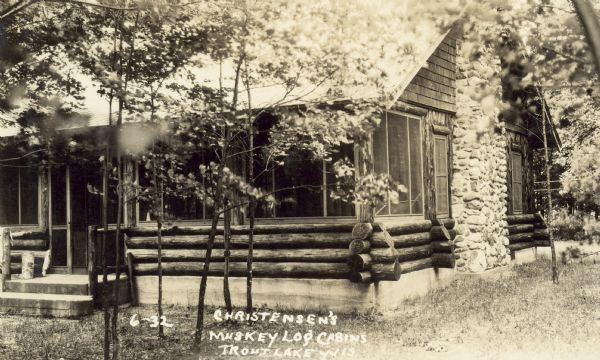 Caption reads: "Christensen's Muskey Log Cabins, Trout Lake, Wis." Steps lead up to a door to the porch on the left. There is a stone chimney on the right side of the cabin.