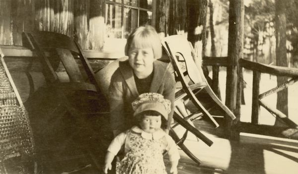 A girl (Marguerite Christensen) is standing on a porch leaning over a doll she is holding in front of her. The chairs on the porch are leaning against the wall, and a snowshoe is on the left. Rustic-style porch railings are in the background.