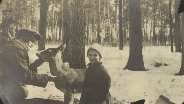 A man is examining or petting a deer while a child is standing nearby. Another man is standing in the background on the left. Snow is on the ground. Caption reads: "John and Pet Deer."