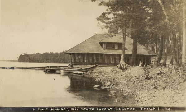 View along shoreline toward a boathouse. Caption on print reads: "Boat House: Wis. State Forest Reserve. Trout Lake."