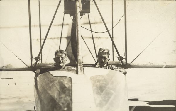 Caption reads: "Jack Vilas & Weaver." View along front of seaplane, with two men sitting inside. Both men have goggles. Water is in the background.