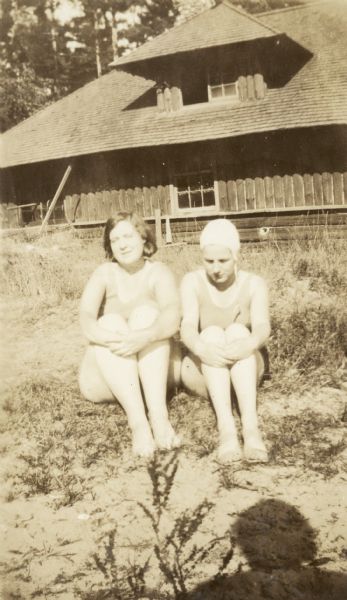Two women are sitting on the ground outdoors, wearing bathing suits. The woman on the right is wearing a bathing cap. A large log building in the background is the boathouse.