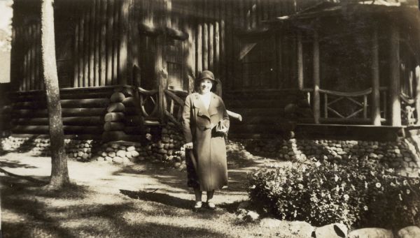 A woman wearing a long coat and hat, and holding a handbag is standing in front of a lodge.