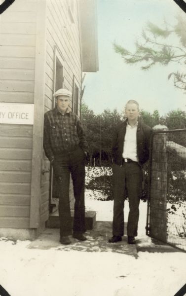 Outdoor portrait of two men posing next to a building with snow on the ground. The man on the left is leaning against the side of a building, which has a sign that reads, in part: "ry office." The other man on the right is near a fence post. Caption in album reads: "Hans & Barney."