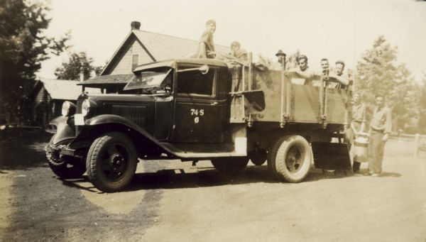 Group of men in a truck, with 74-S on the driver side door. Two men are standing behind the truck holding a milk can. Buildings are in the background. Caption in album reads: "Some boys from White Sand Camp."