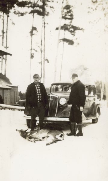 Two men are standing with a dead deer in front of an automobile. The corner of a building is in the background on the right. Caption in album reads: "Phil & Frank Long, Game Wardens with 2 confiscated deer."