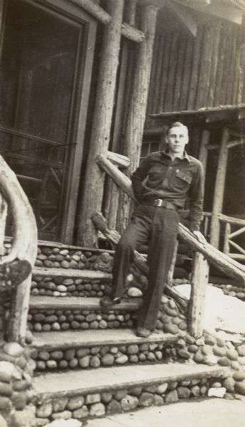 Caption in album reads: "John." A man wearing a long sleeved shirt and long pants is standing and leaning on the railing of a rustic building.