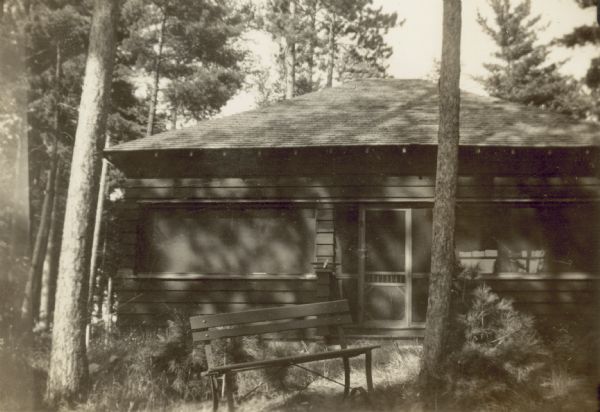Caption in album reads: "The Harrington Cottage." The cottage has a door between two large windows, and is surrounded by pine trees. There is a bench in the grass in the foreground.