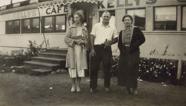 Two women and a man are standing in front of Kelly's Cafe. There is an awning in front of the entrance. The man in the center is holding a bottle in one hand and a cigarette in the other. Caption in album reads: "Mary, Kelley, & Dott."