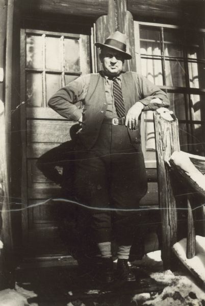 Portrait of a man standing on snowy steps in front of the door of a building. He is resting one elbow on the post of a railing.