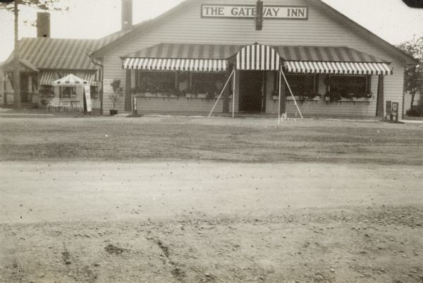 View across unpaved road towards The Gateway Inn. There are striped awnings over the windows, and on the left are chairs near a table with an umbrella. Caption in album reads: "The 'Gateway' at Land o' Lakes."