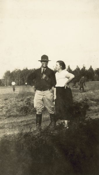 A man and woman are standing together outdoors. People are in a field in the background. Written on back: "Al Peterson (Lieutenant)."