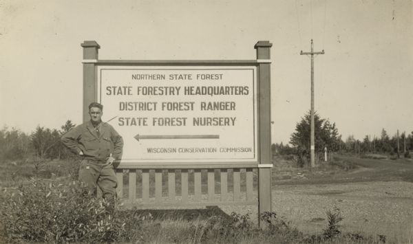A man is standing outdoors next to a sign that reads: "Northern State Forest, State Forest Headquarters, District Forest Ranger, State Forest Nursery. Wisconsin Conservation Commission."