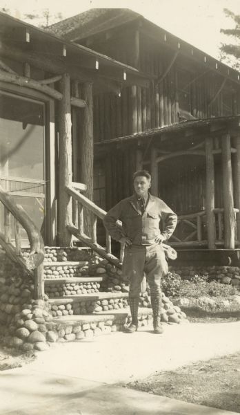 Man standing on the sidewalk outdoors in front of a building constructed of logs, with rustic railings, and stone slab steps with rock detail on the stairs leading up to the screen door.