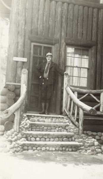 A man wearing a coat, necktie and cap is standing at the top of wood and slab stone steps with rustic railing at an entrance to the lodge at Trout Lake. A sign to the left of the door reads: "Office."